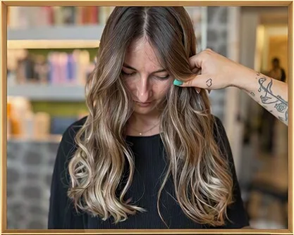 Una cliente con lunghe onde balayage castane e bionde; una mano tatuata solleva una ciocca vicino al viso.