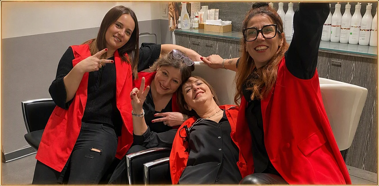 Quattro ragazze dello staff in gilet rosso sulle poltrone del lavaggio, sorridenti, una fa il segno della vittoria e un'altra scatta un selfie.